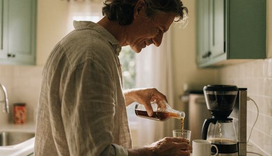 Man in a bright kitchen pouring a small morning serving of Haelan 951 fermented soy beverage while coffee brews