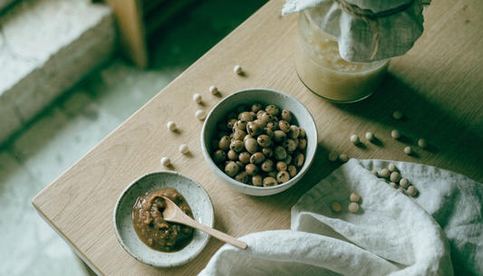 Fermented soy foods on a light oak table in soft natural light: a ceramic bowl of fermented soybeans, a dish of dark miso paste with a wooden spoon, a cloth-covered fermentation jar, and scattered dried soybeans.