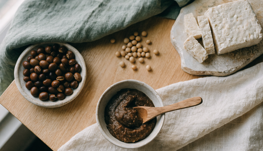 Overhead flat lay of traditional fermented soy foods on light oak: a ceramic bowl of dark miso paste, sliced tempeh on a stone board, cooked soybeans and scattered dried soybeans on cream linen