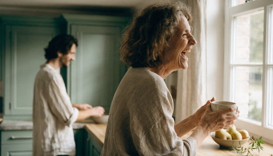A woman in her fifties laughing with a mug of tea in a sunlit autumn kitchen while a younger man reaches for a bowl nearby, illustrating a fall wellness reset routine