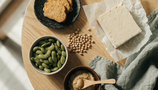 Overhead flat lay of soy protein foods on a light oak table: sliced tempeh, shelled edamame, a block of tofu, a bowl of miso paste and dried soybeans