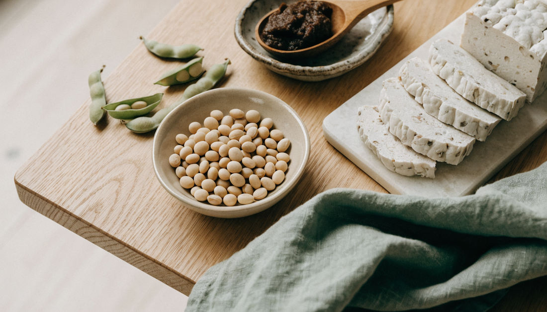 Flat lay of fermented soy foods on a light oak table: a bowl of whole soybeans, dark miso paste in a stoneware dish, sliced tempeh on a stone board, and fresh soybean pods on a sage green linen napkin
