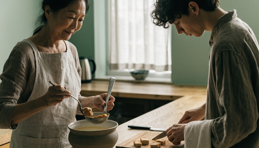 A mother and adult son cooking with fermented foods at home, stirring miso into soup off the heat beside pan-seared tempeh.