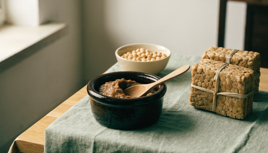 Doenjang, Korean fermented soybean paste, in an earthenware crock beside dried meju blocks and soybeans on a light oak table.
