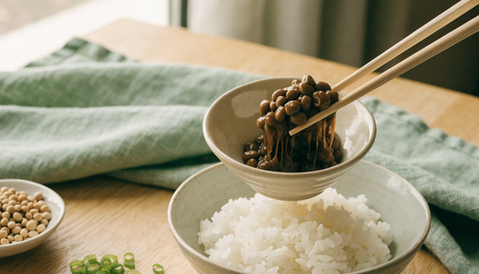 A ceramic bowl of natto, Japanese fermented soybeans with sticky strands, lifted with chopsticks over rice on a light oak table.