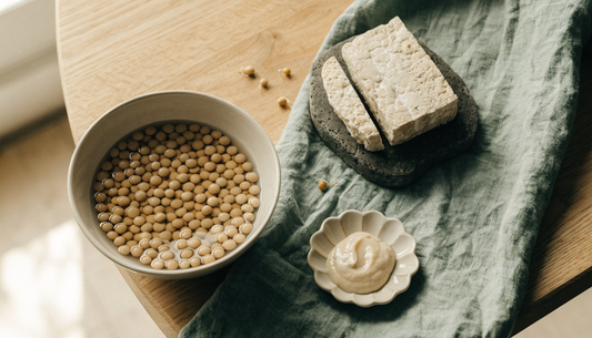 Soybeans soaking in a ceramic bowl beside sliced tempeh, miso paste and sprouted soybeans on a light oak table, illustrating how fermentation and soaking affect phytic acid in soy.